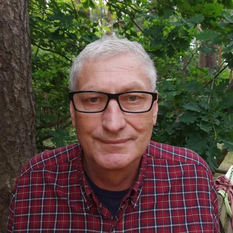 Doświadczony psychoterapeuta, ubrany w bordową koszulę w kratę, stoi w terenie lesistym.